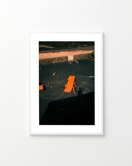The Short Romantic Story Between a Chair and a Shadow Poster features three orange lounge chairs creating captivating shadows on black sand under bright light, with two more chairs by a white fence in the background, all viewed from an elevated angle on a tranquil beach.