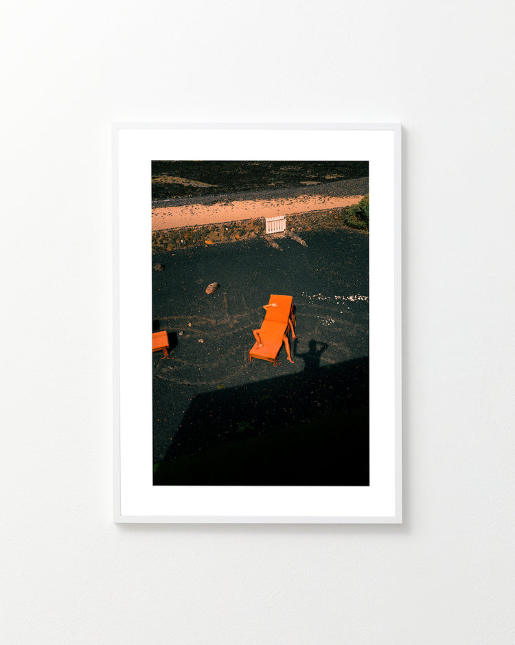 The Short Romantic Story Between a Chair and a Shadow Poster features three orange lounge chairs creating captivating shadows on black sand under bright light, with two more chairs by a white fence in the background, all viewed from an elevated angle on a tranquil beach.