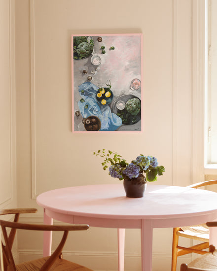A pink dining table with a black vase of purple and green flowers is paired with a wooden chair. Above, the We Taste Better Cooked poster featuring artichokes, lemons, and a tea towel adds culinary artistry to the light-colored wall.