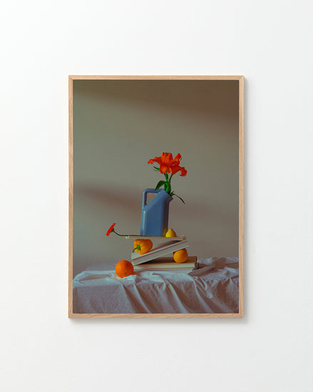 The Hanging in There Poster features a vibrant scene: a blue watering can with an orange flower on open books, surrounded by two oranges and a lemon on white cloth, evoking Berlins artistic charm against a simple backdrop.
