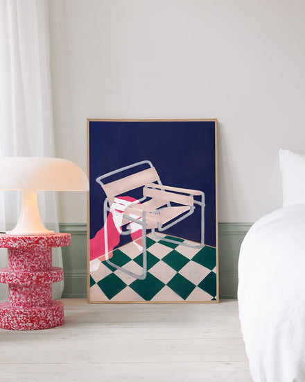 A minimalist room showcases the Bauhaus 02 Poster with a framed abstract of a white Wassily chair over a red and green checkered floor, set against a dark blue backdrop. The artwork leans beside a modern lamp on a bold red table, capturing Bauhaus modernisms essence.