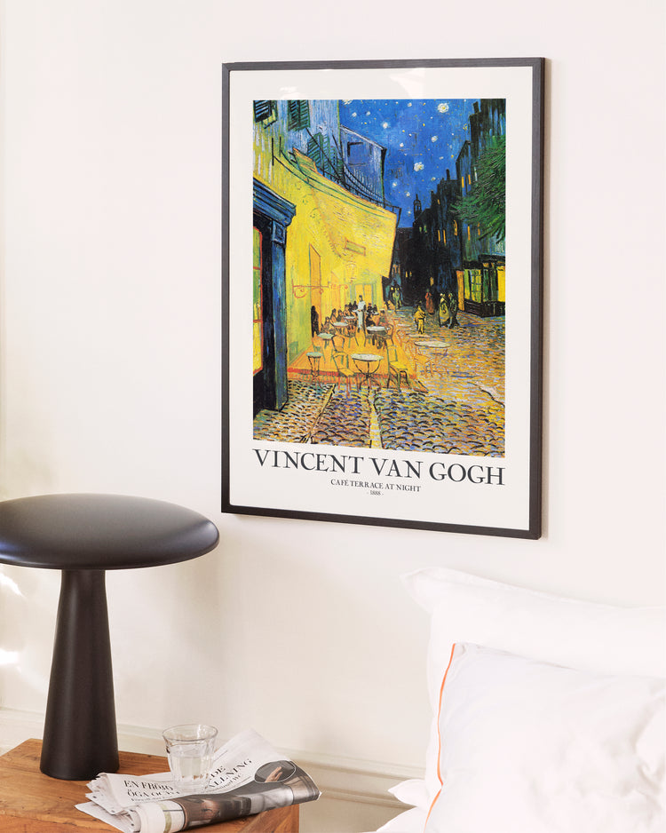 Café Terrace at Night by Vincent van Gogh Poster framed on a white wall, depicting a vibrant café scene under a starry evening sky. Below it, the artists name and title are displayed. Nearby, a small bedside table holds magazines and a glass.