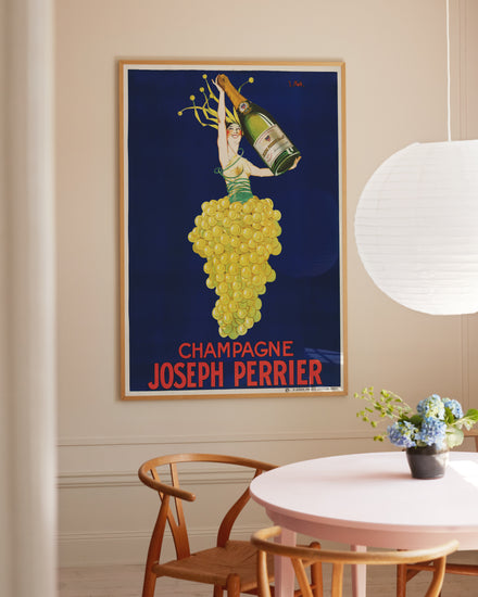 The room features a Champagne Joseph Perrier Poster framed on the wall, depicting a grape-made figure holding a champagne bottle against a blue backdrop, complemented by a wooden chair, round table with blue flowers in a vase, and a hanging white paper lantern.