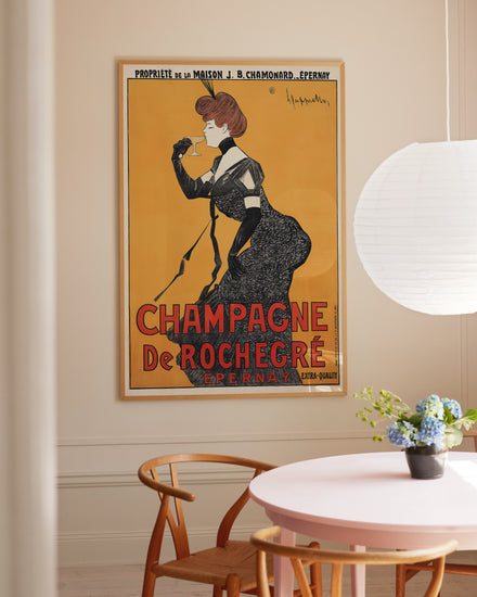 The Champagne de Rochegré Epernay vintage poster, exuding Art Deco charm, shows a woman in a black dress sipping wine. Its framed on a beige wall near a round pink table with blue flowers beneath a white paper lantern.