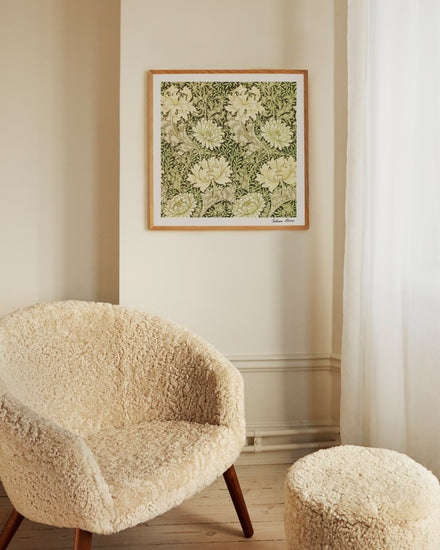 A cozy room features a textured armchair and footstool with wooden legs. The wall showcases the Chrysanthemum Pattern by William Morris poster. Soft light through white curtains enhances the warm and inviting ambiance.