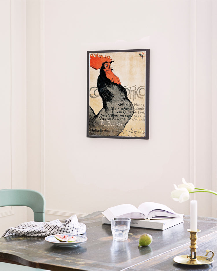 A Cocorico by Théophile-Alexandre Steinlen poster adds vintage charm to the wall. Below, a wooden table holds an open book, a glass of water, and a black-patterned white napkin. In front, a white tulip in a glass vase embodies French Art Deco elegance.