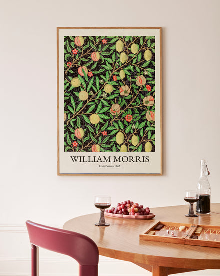 A wooden dining table holds two glasses of red wine, a bowl of grapes, and a backgammon set. Above it is a vibrant Fruit Pattern by William Morris Poster adorning the white wall.