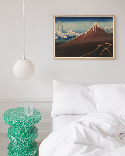 A minimalist bedroom has a white bed with white bedding, a green speckled side table with a glass, and a round white pendant light. A blue and red toned framed poster titled Fuji above the Lightning by Katsushika Hokusai hangs above.