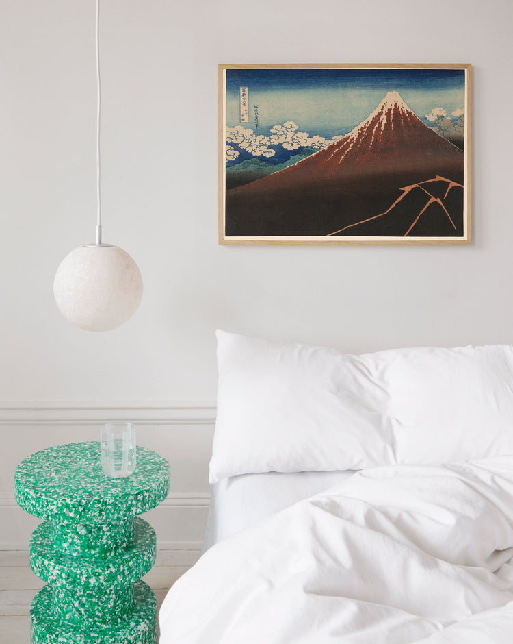 A minimalist bedroom has a white bed with white bedding, a green speckled side table with a glass, and a round white pendant light. A blue and red toned framed poster titled Fuji above the Lightning by Katsushika Hokusai hangs above.