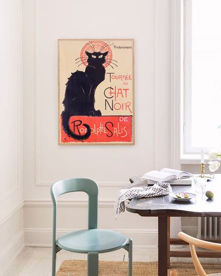 A room styled with Parisian chic includes a green chair, a small white table with books and a bowl, and a Le Chat Noir by Théophile-Alexandre Steinlen poster featuring a black cat. The space has white walls and minimal decor.