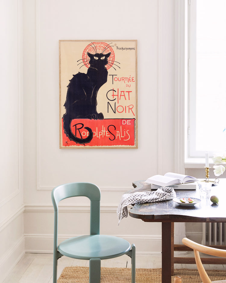 A room styled with Parisian chic includes a green chair, a small white table with books and a bowl, and a Le Chat Noir by Théophile-Alexandre Steinlen poster featuring a black cat. The space has white walls and minimal decor.