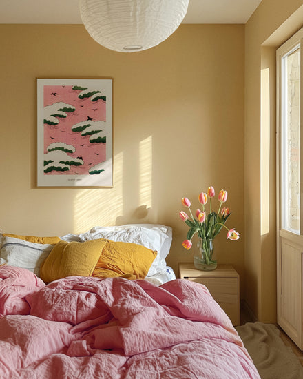 A cozy bedroom features a bed with dreamy pink sky and yellow bedding. A wooden nightstand holds a tulip vase, while the Pink Sky by Watanabe Seitei poster, inspired by Japanese woodblock prints, adorns the wall. Sunlight pours through a large window creating patterns on the wall.