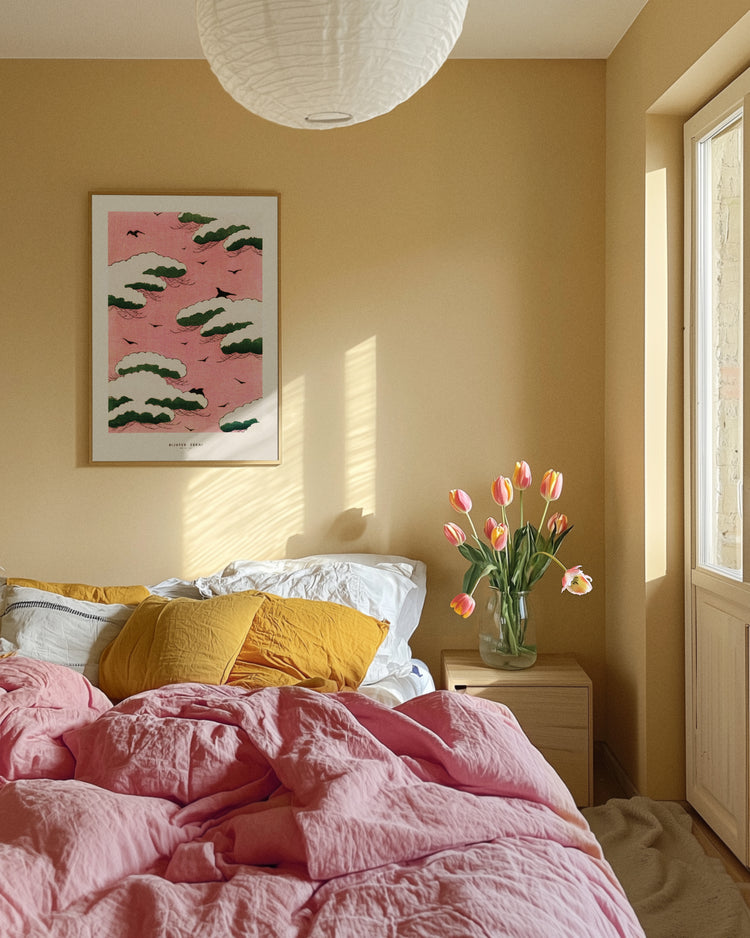 A cozy bedroom features a bed with dreamy pink sky and yellow bedding. A wooden nightstand holds a tulip vase, while the Pink Sky by Watanabe Seitei poster, inspired by Japanese woodblock prints, adorns the wall. Sunlight pours through a large window creating patterns on the wall.