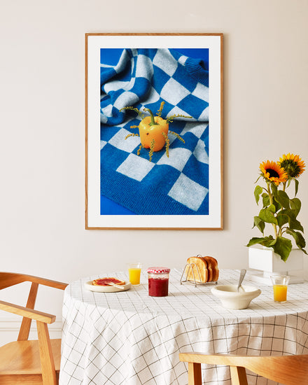 A dining area with a checked tablecloth and wooden chairs showcases juice, bread, and jam. The playful touch comes from the Little Visitor Poster, depicting a bell pepper as the sun on a checkered cloth. A sunflower plant completes this bright and inviting setting.