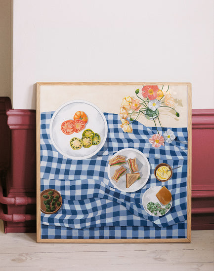 The Poppies and Picnics Poster features a painting of a blue and white checkered tablecloth with sliced tomatoes, sandwiches, and leafy greens. A vibrant floral arrangement completes the summer romance scene. Perfect for adding charm to your kitchen wall, it leans gracefully against a rustic backdrop.