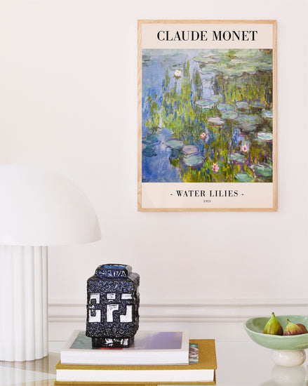 A soothing interior showcases a Claude Monet Water Lilies by Claude Monet 1915 Poster, elegantly framed in light wood. A white lamp and a small decorative box sit on stacked books, while figs in a bowl add nature to this French Impressionism-inspired scene.