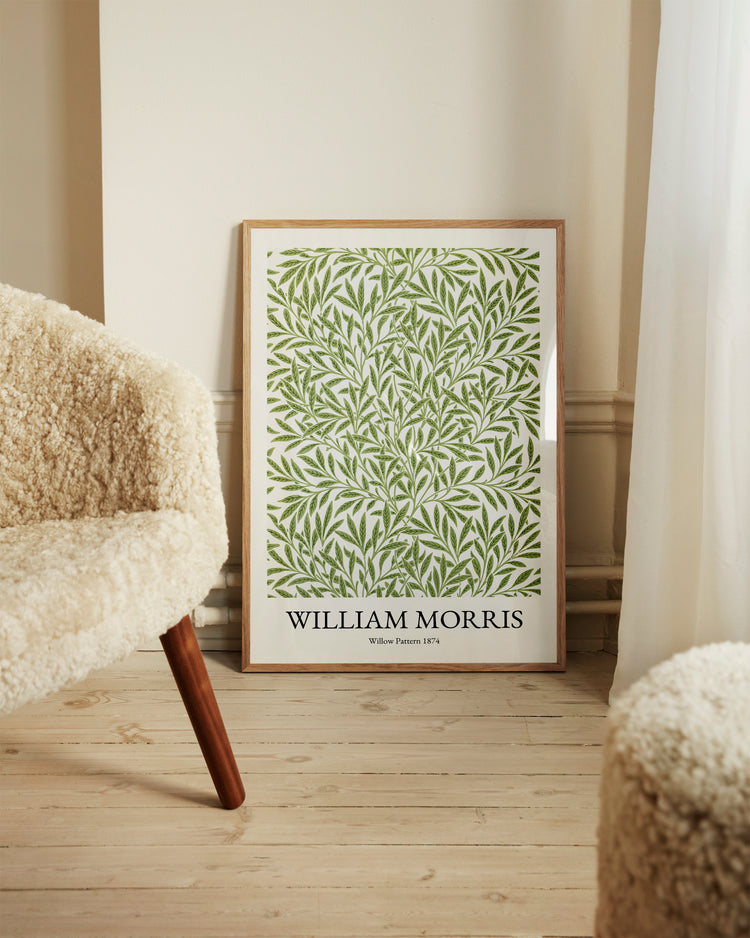 A framed William Morris Pattern 1874 Poster with a green leaf design leans against a wall. Beside it, a cream-colored textured chair is placed on a light wooden floor, bathed in soft natural light through the curtain, enhancing the scenes Victorian charm.