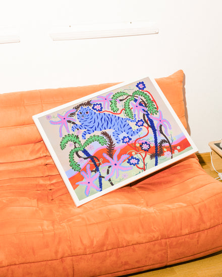 The Dizzy Daizy Poster, a vibrant abstract piece with tropical plants and flowers, leans against an orange sofas backrest. The couch faces a surreal landscape on a plain white wall and sits elegantly on the wooden floor.