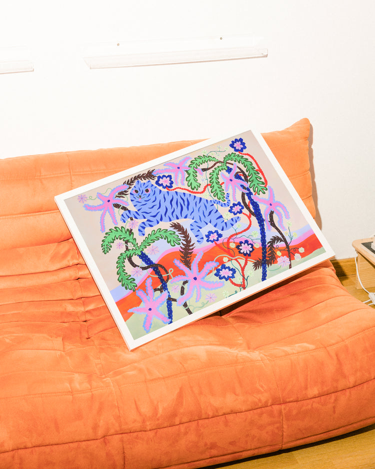 The Dizzy Daizy Poster, a vibrant abstract piece with tropical plants and flowers, leans against an orange sofas backrest. The couch faces a surreal landscape on a plain white wall and sits elegantly on the wooden floor.