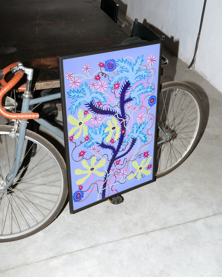 A bicycle with brown handlebars leans against a wall, while the vibrant Someplace Both Wonderful & Strange poster featuring blue, yellow, and pink elements is propped against its frame on a concrete floor, enhancing the indoor ambiance.