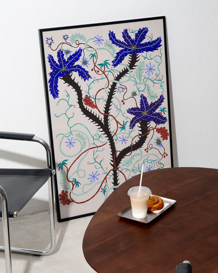 The Wild Whirling Cherries Poster leans against the white wall. Nearby, a black chair complements a round wooden table holding a tray with a drink and pastry, creating a lively scene reminiscent of wildflowers blooming in this tranquil corner.