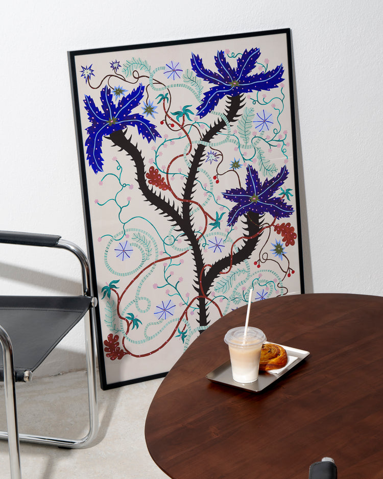 The Wild Whirling Cherries Poster leans against the white wall. Nearby, a black chair complements a round wooden table holding a tray with a drink and pastry, creating a lively scene reminiscent of wildflowers blooming in this tranquil corner.