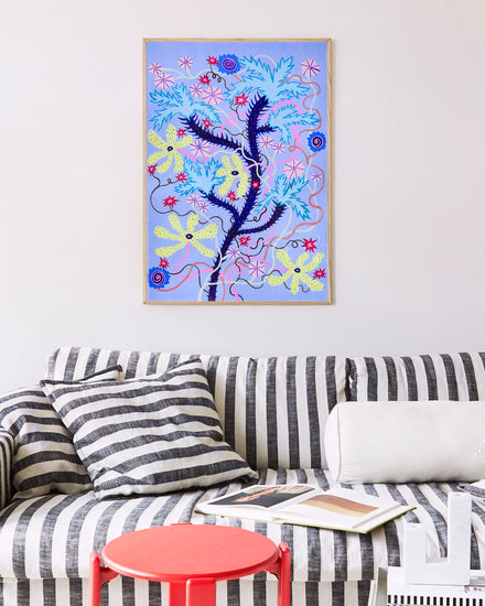 A striped couch with white and black cushions is in front of a white wall featuring Yoyos signature Someplace Both Wonderful & Strange poster, showcasing wildflowers against a dreamy blue background. A small red side table with an open book adds charm.