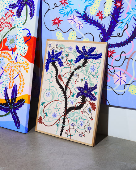The Wild Whirling Cherries Poster features abstract art with intertwining black branches, blue leaves, and wildflowers amid colorful swirls. It leans against a wall on a gray floor with vibrant, similarly styled artworks.