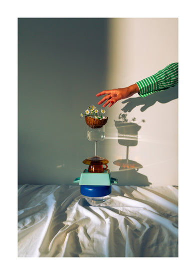 Small bowls, a wine glass delicately balanced atop each other, and at the top, a half coconut adorned with flowers. A hand reaching in for the blooms. White border.