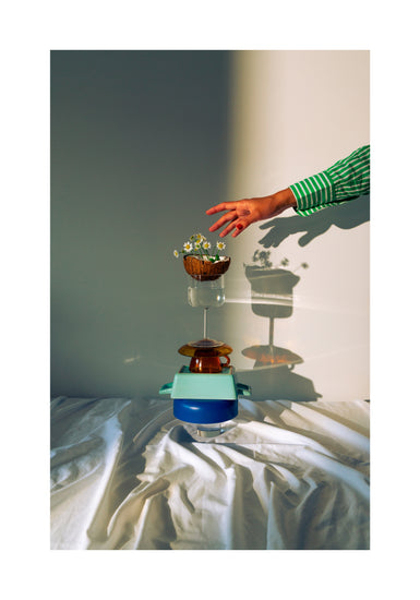 Small bowls, a wine glass delicately balanced atop each other, and at the top, a half coconut adorned with flowers. A hand reaching in for the blooms. White border.