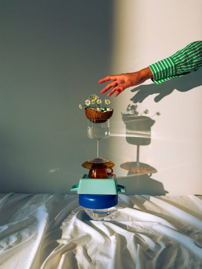 Small bowls, a wine glass delicately balanced atop each other, and at the top, a half coconut adorned with flowers. A hand reaching in for the blooms.