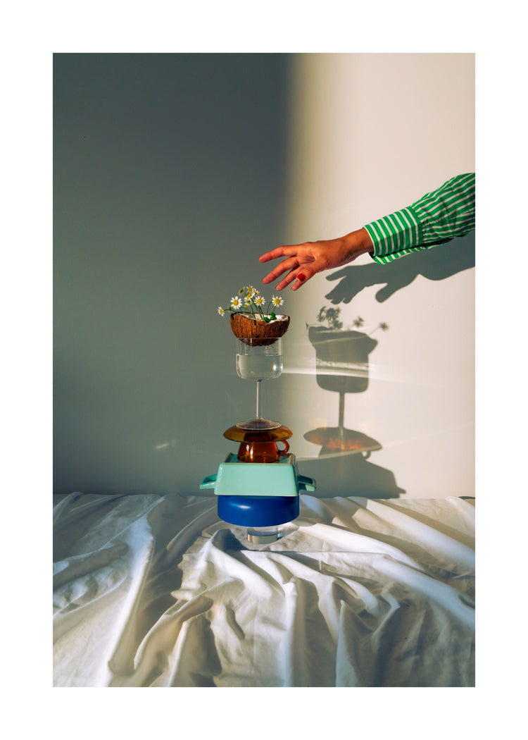 Small bowls, a wine glass delicately balanced atop each other, and at the top, a half coconut adorned with flowers. A hand reaching in for the blooms. White border.