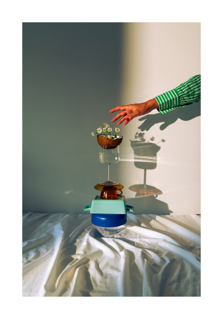 Small bowls, a wine glass delicately balanced atop each other, and at the top, a half coconut adorned with flowers. A hand reaching in for the blooms. White border.