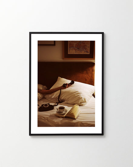 The Do Not Disturb Poster showcases a cozy bed scene, complete with white pillows and sheets. A hand holds an old-school phone, while nearby an open book and cup of coffee rest under dim, warm lighting, exuding retro photography charm.