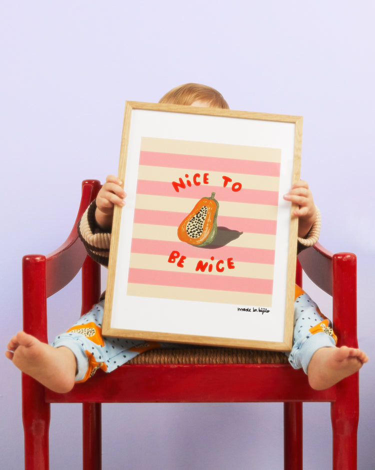 A baby sitting on a red chair, holding up a poster of a papaya in an oak frame in front of them.