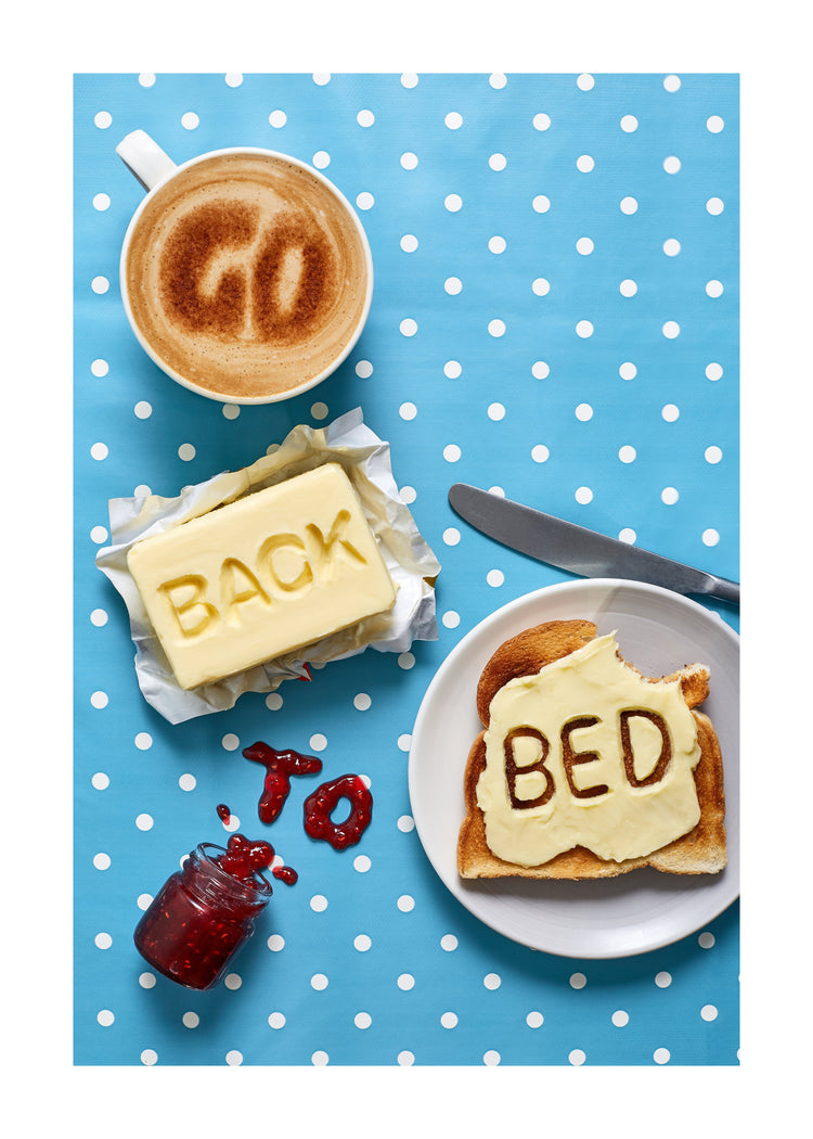 A mug of coffee spells 'GO' in froth, 'BACK' is intricately etched in butter, 'TO' is crafted from fruity jam, and 'bed' is toasted onto cheese toast. All arranged on a delightful blue tablecloth adorned with white polka dots.