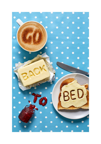 A mug of coffee spells 'GO' in froth, 'BACK' is intricately etched in butter, 'TO' is crafted from fruity jam, and 'bed' is toasted onto cheese toast. All arranged on a delightful blue tablecloth adorned with white polka dots.
