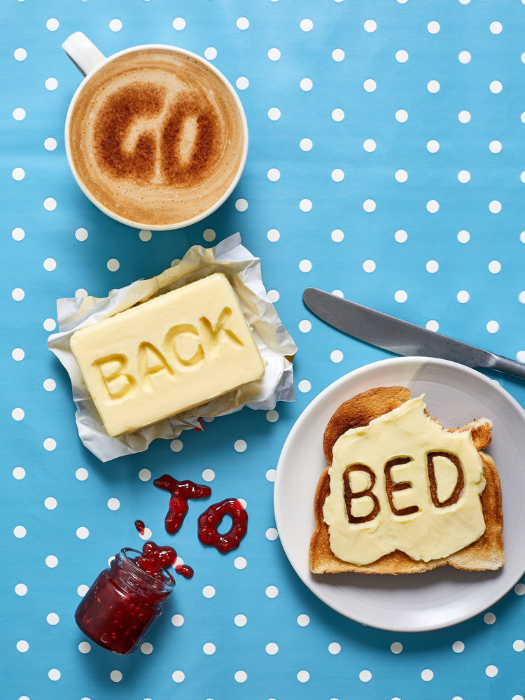 A mug of coffee spells 'GO' in froth, 'BACK' is intricately etched in butter, 'TO' is crafted from fruity jam, and 'bed' is toasted onto cheese toast. All arranged on a delightful blue tablecloth adorned with white polka dots.