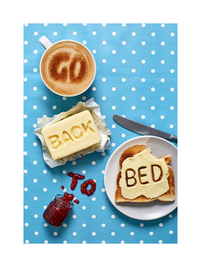 A mug of coffee spells 'GO' in froth, 'BACK' is intricately etched in butter, 'TO' is crafted from fruity jam, and 'bed' is toasted onto cheese toast. All arranged on a delightful blue tablecloth adorned with white polka dots.
