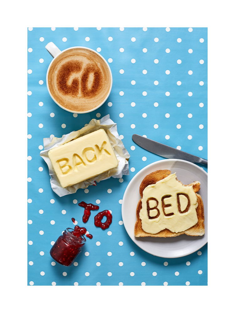 A mug of coffee spells 'GO' in froth, 'BACK' is intricately etched in butter, 'TO' is crafted from fruity jam, and 'bed' is toasted onto cheese toast. All arranged on a delightful blue tablecloth adorned with white polka dots.