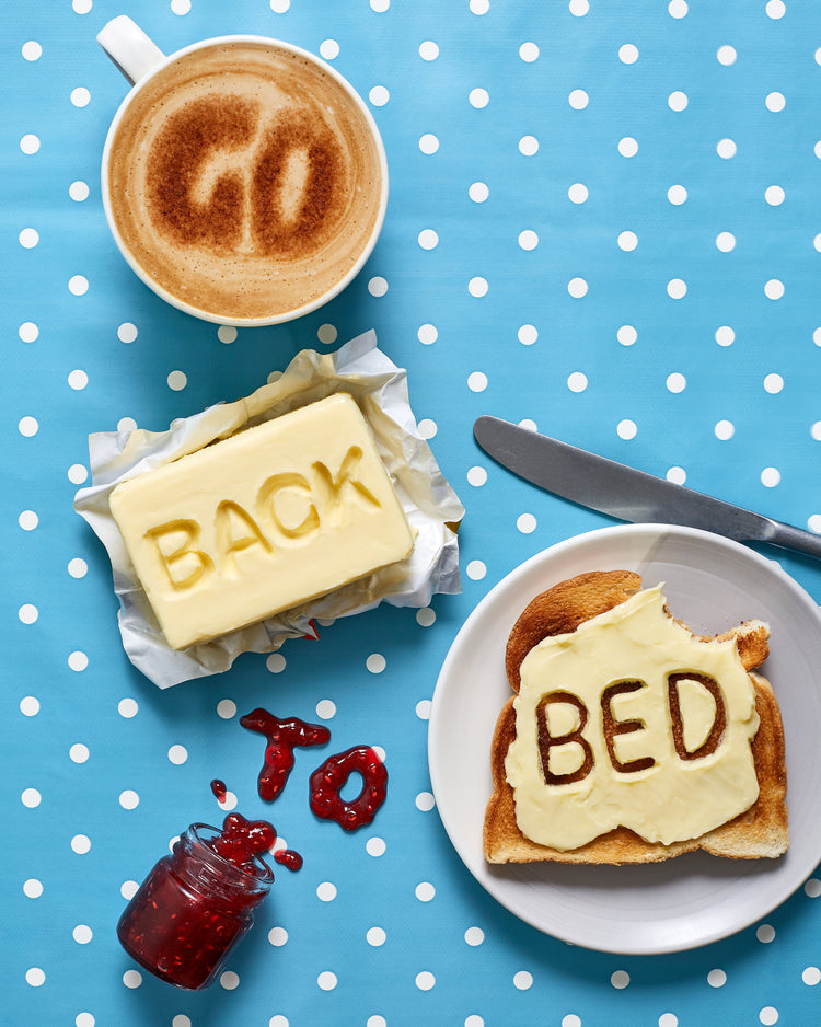A mug of coffee spells 'GO' in froth, 'BACK' is intricately etched in butter, 'TO' is crafted from fruity jam, and 'bed' is toasted onto cheese toast. All arranged on a delightful blue tablecloth adorned with white polka dots.
