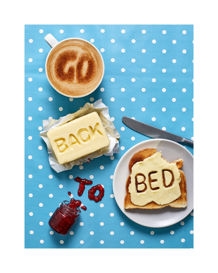 A mug of coffee spells 'GO' in froth, 'BACK' is intricately etched in butter, 'TO' is crafted from fruity jam, and 'bed' is toasted onto cheese toast. All arranged on a delightful blue tablecloth adorned with white polka dots.