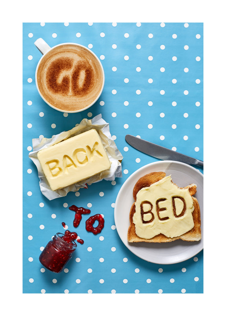 A mug of coffee spells 'GO' in froth, 'BACK' is intricately etched in butter, 'TO' is crafted from fruity jam, and 'bed' is toasted onto cheese toast. All arranged on a delightful blue tablecloth adorned with white polka dots.