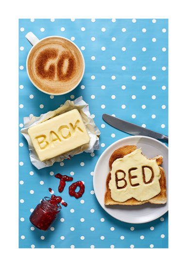 A mug of coffee spells 'GO' in froth, 'BACK' is intricately etched in butter, 'TO' is crafted from fruity jam, and 'bed' is toasted onto cheese toast. All arranged on a delightful blue tablecloth adorned with white polka dots.