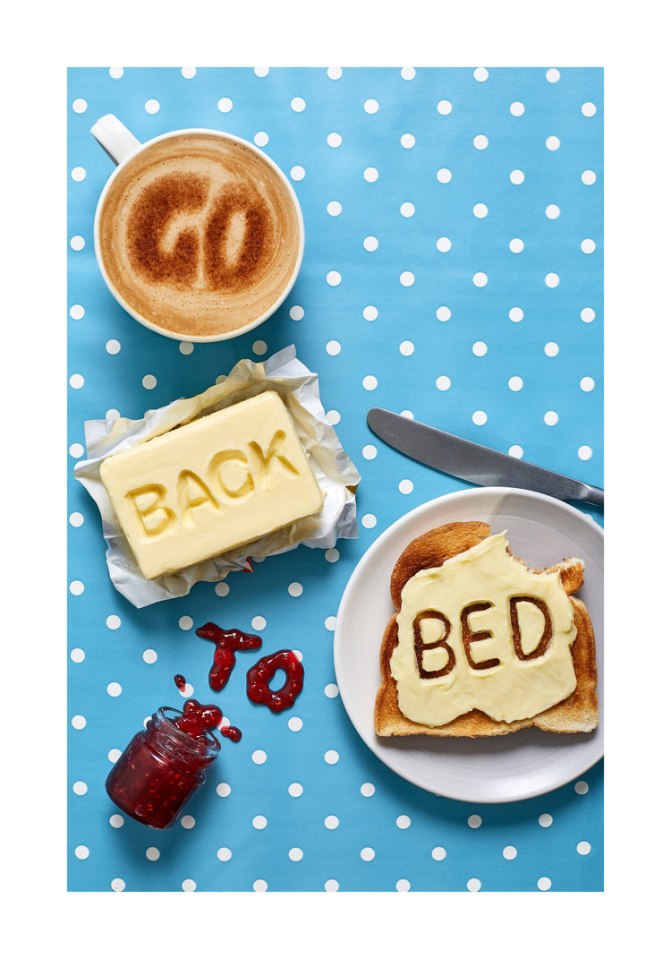 A mug of coffee spells 'GO' in froth, 'BACK' is intricately etched in butter, 'TO' is crafted from fruity jam, and 'bed' is toasted onto cheese toast. All arranged on a delightful blue tablecloth adorned with white polka dots.