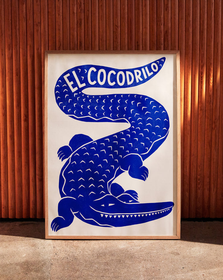 Framed El Cocodrilo Poster by Manne Jalilian features a stylized blue crocodile on a white background with El Cocodrilo elegantly inscribed above, set against a vertical wooden panel wall for added character.