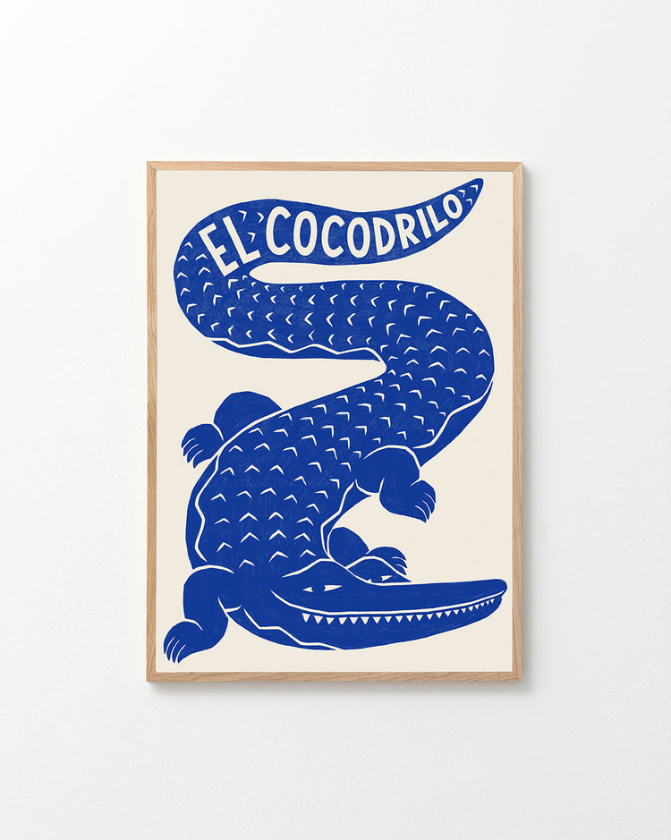 Framed poster with an illustration of a blue crocodile with the text El Cocodrilo is hanging on a white wall. Illustration created by the artist Manne Jalilian.