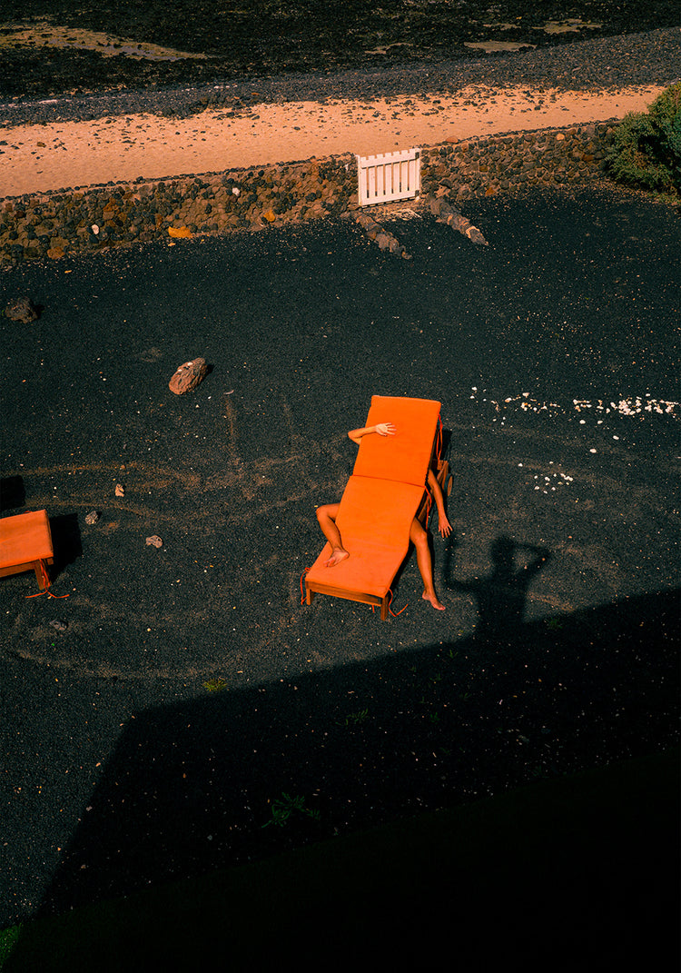 Amidst a rocky landscape, an outstretched arm peeks from behind an orange bench as shadows play on the ground, including that of a photographer. Its reminiscent of The Short Romantic Story Between a Chair and a Shadow Poster.