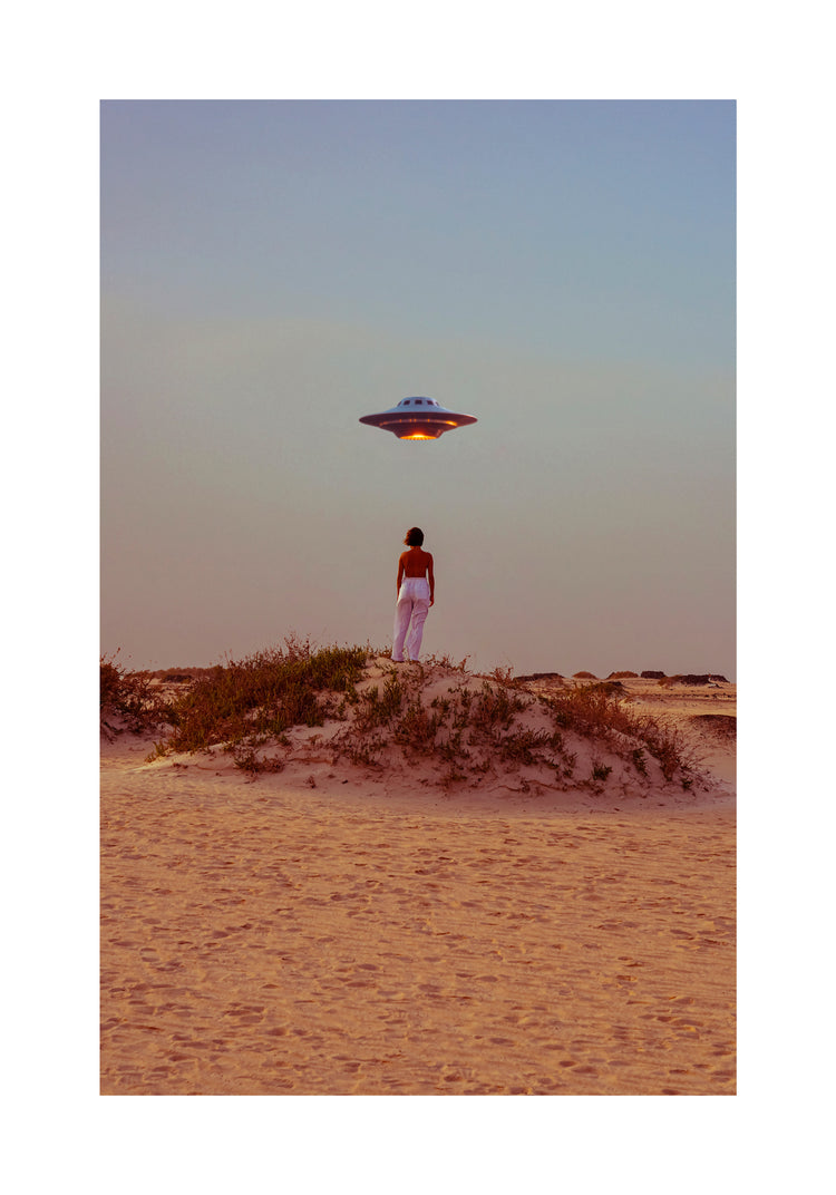 On a sandy desert dune, an individual captures a self-portrait gazing at a spaceship against the clear sky. The scene is lit warmly, adding to the surreal atmosphere depicted in the Hey Luke, our Uber is Here! Poster.