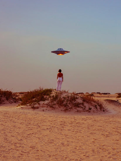 A person in white pants stands on a deserts sandy terrain, taking a selfie with the Hey Luke, our Uber is Here! Poster showing a UFO in the clear dusk sky. Sparse bushes decorate the orange-hued horizon.